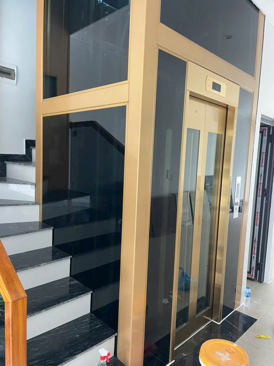 Thang máy gia đình khung kính cửa màu vàng đặt cạnh cầu thang trong nhà